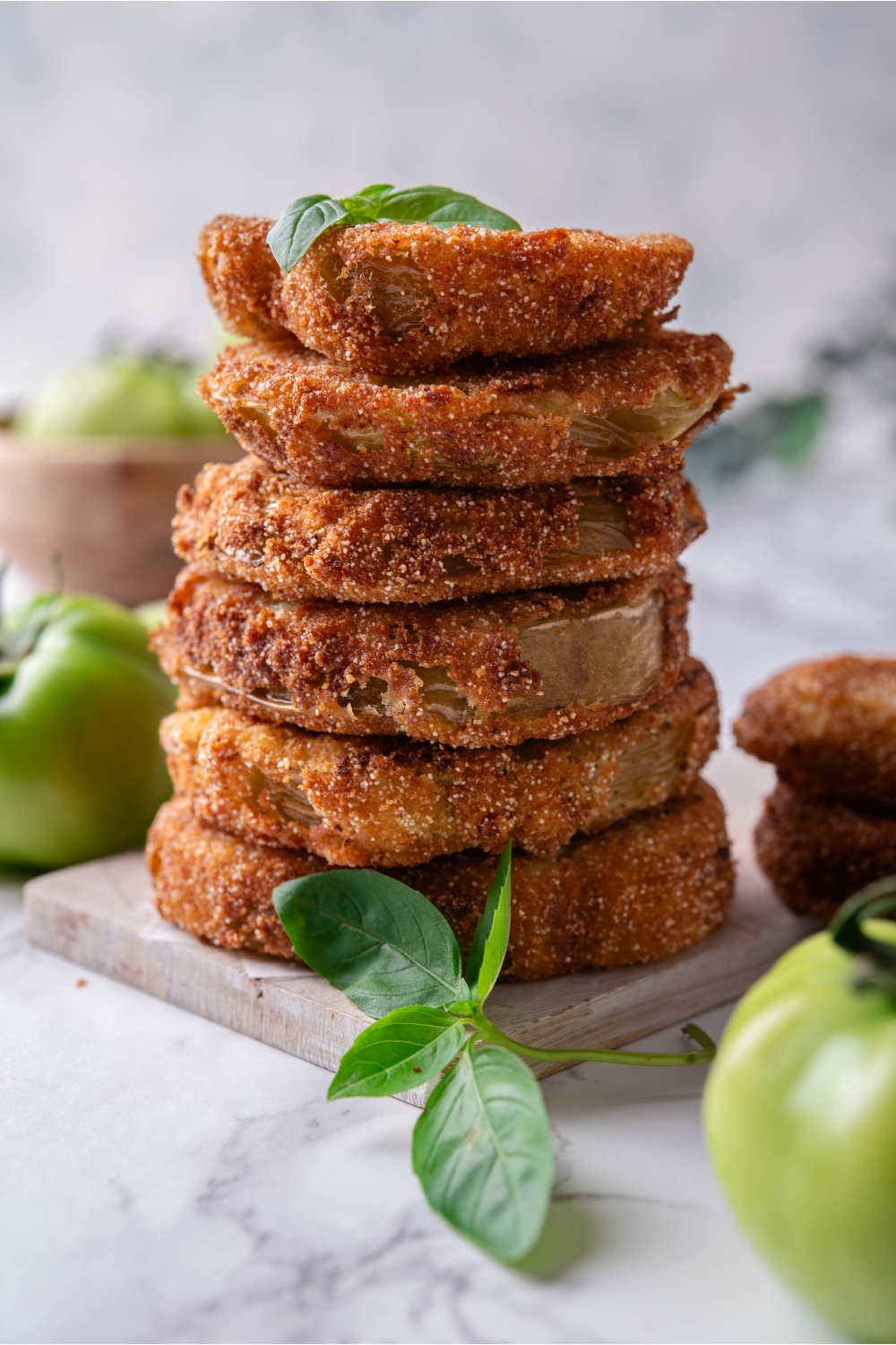 Six breaded and fried green tomatoes stacked evenly on top of each other with a garnish of green herbs on top and fresh green tomatoes surrounding the stack.