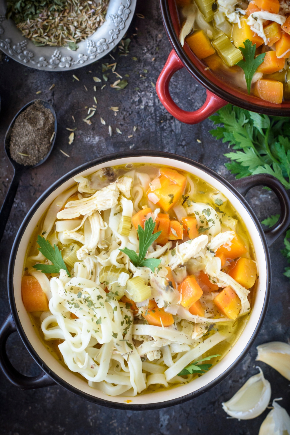 A bowl of chicken noodle soup with chunks of carrot, fresh herbs, and dried herbs sprinkled on top.