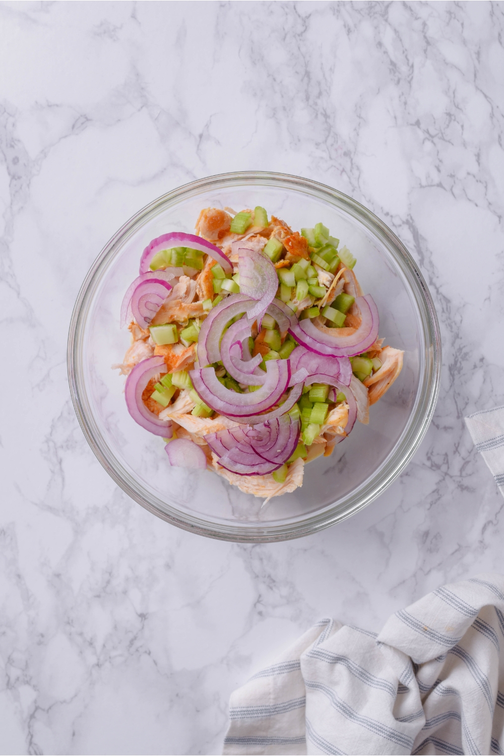 A clear bowl filled with shredded seasoned chicken, thinly sliced red onion, and diced celery.