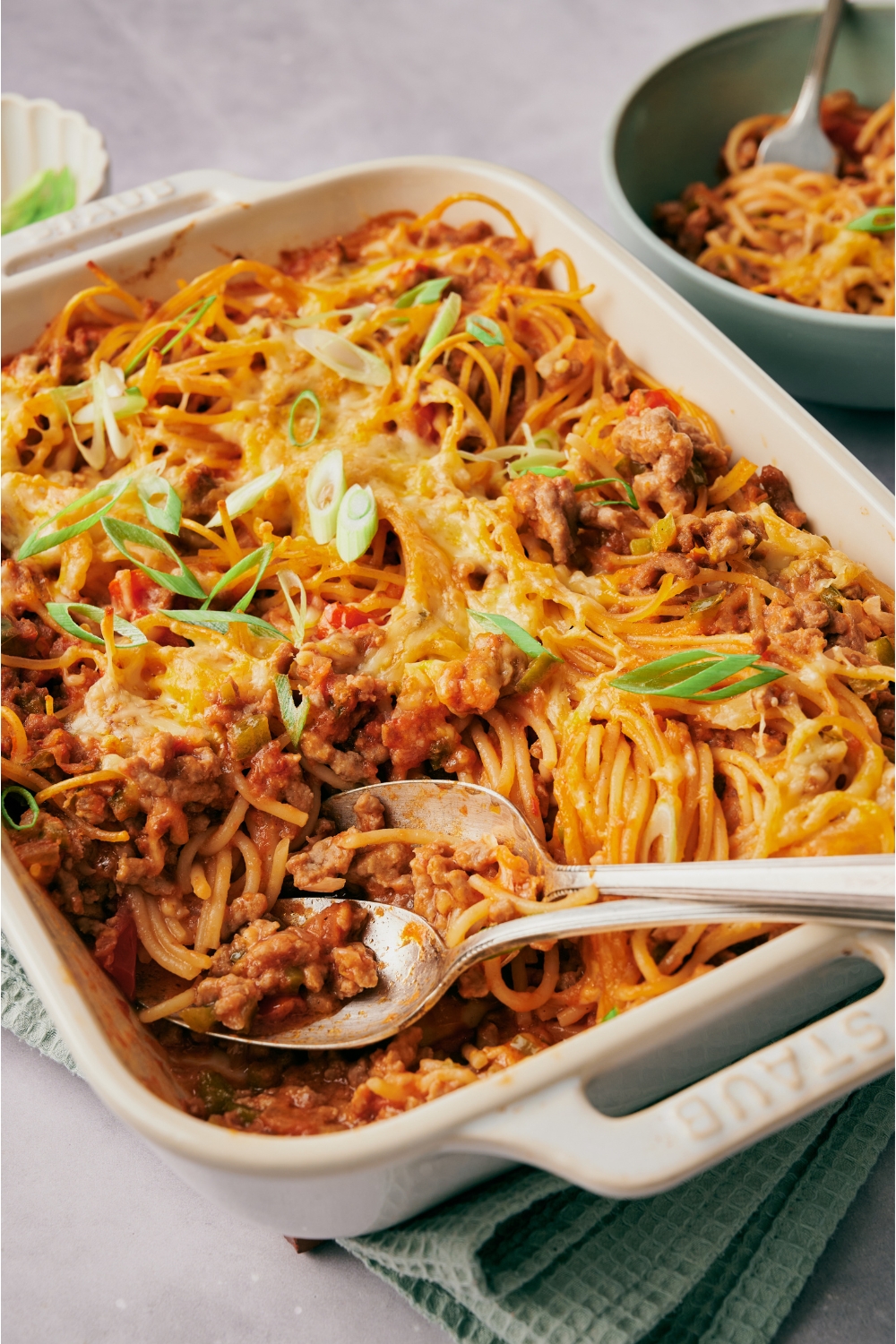 A white baking dish filled with cooked spaghetti casserole with one serving being scooped out using two spoons.