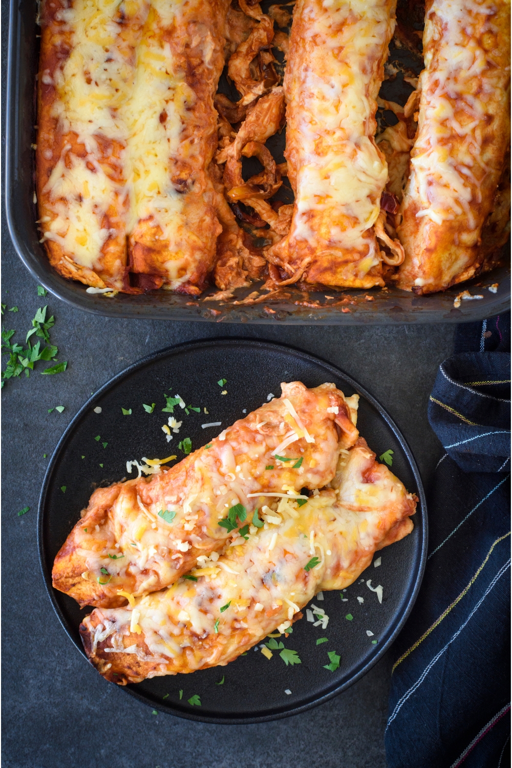 Two enchiladas on a black plate garnished with fresh herbs next to a baking dish containing the rest of the enchiladas.