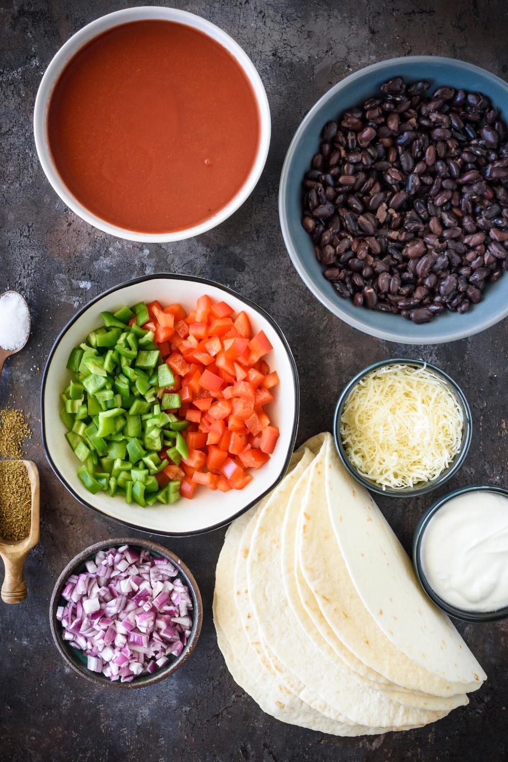 A countertop with multiple bowls with separate ingredients like peppers, onions, beans, cheese, sour cream, enchilada sauce, and flour tortillas.