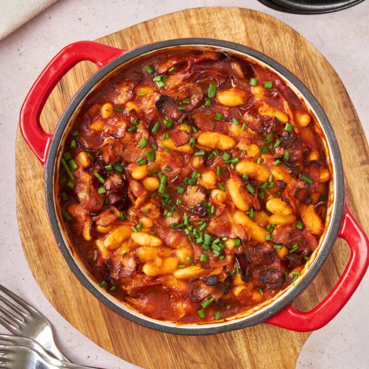 A baked bean casserole in a round baking dish on a wooden cutting board.