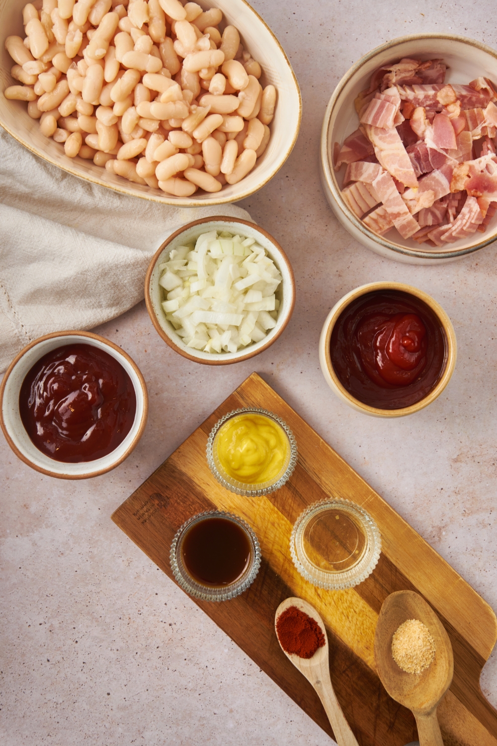 A bowl of great norther beans, a bowl of bacon, a bowl of onion, a bowl of bbq sauce, a bowl of mustard, a bowl of ketchup, a spoon of paprika, and a spoon of garlic powder.