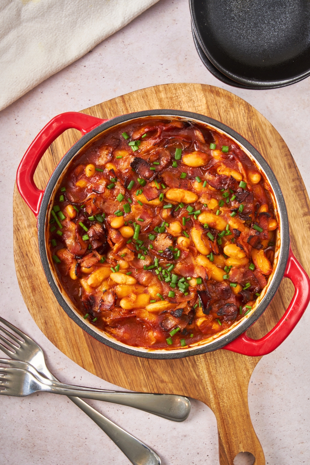 A baked bean casserole in a round baking dish on a wooden cutting board.