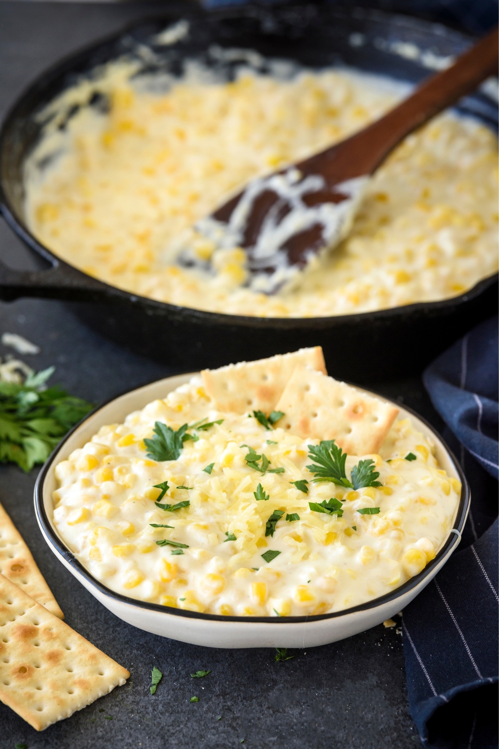 Cream cheese corn in a bowl, garnished with two crackers and some fresh parsley. Behind is a skillet filled with more cream cheese corn and a wooden spatula.