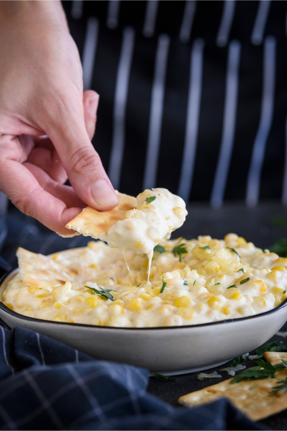 A cracker being dipped into a bowl of cream cheese corn garnished with parsley.