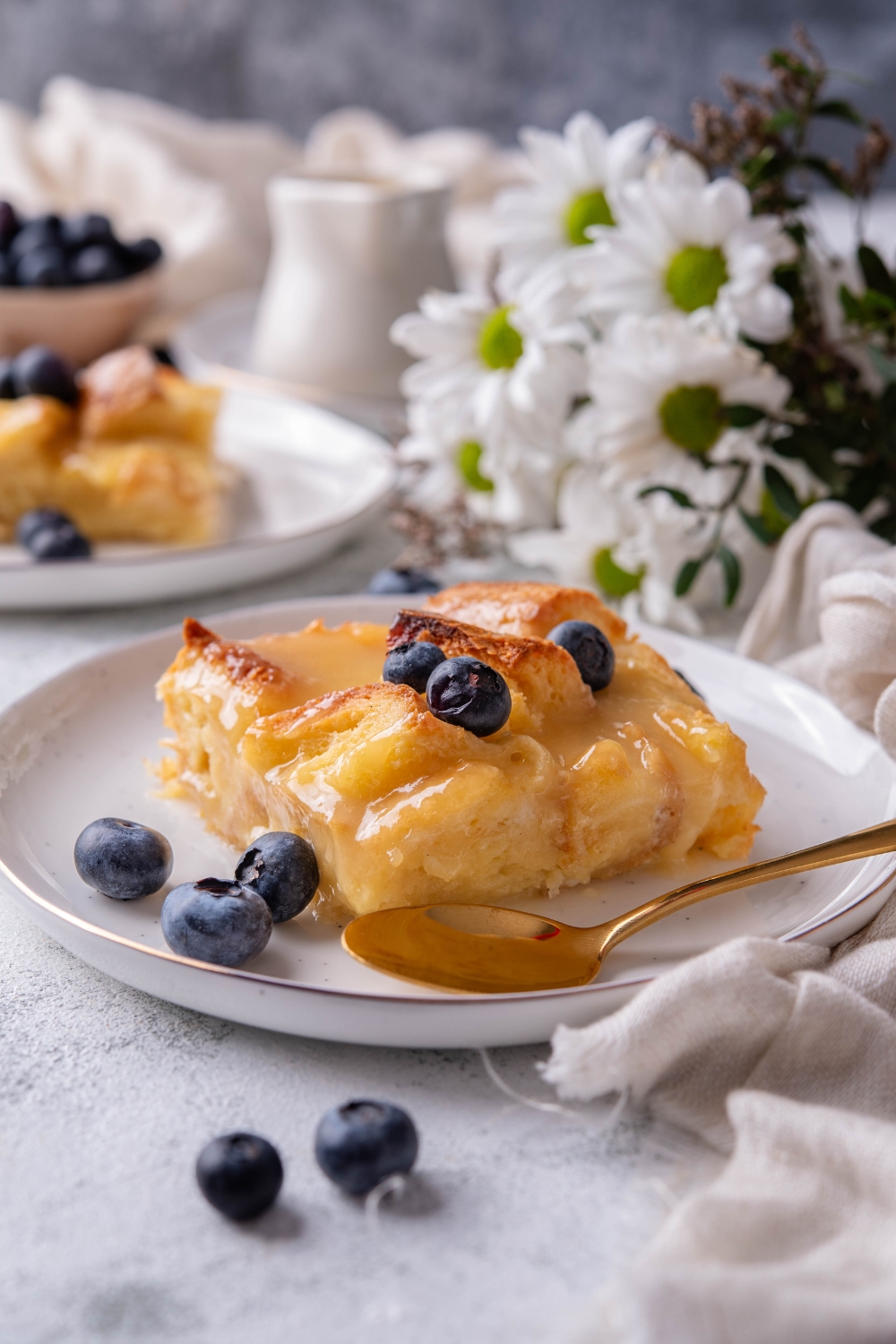 A slice of bread pudding with vanilla sauce and blueberries on a plate, in front of a second plate of bread pudding and blueberries.