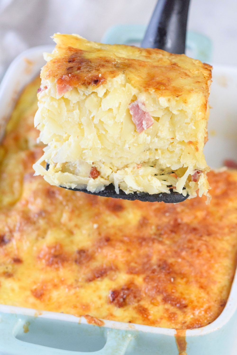 A square of amish breakfast casserole on a black spatula hovering over a casserole dish filled with the casserole..