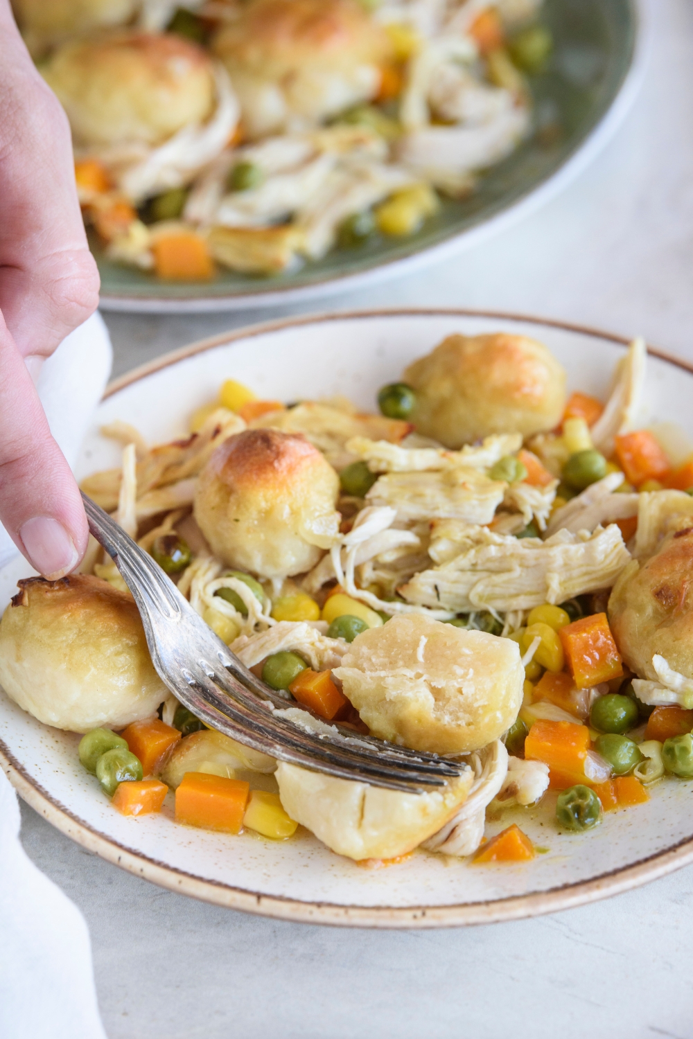A hand holding a fork cutting a dumpling in half. The dumpling is on a plate with peas, carrots, corn, shredded chicken, and more dumplings.