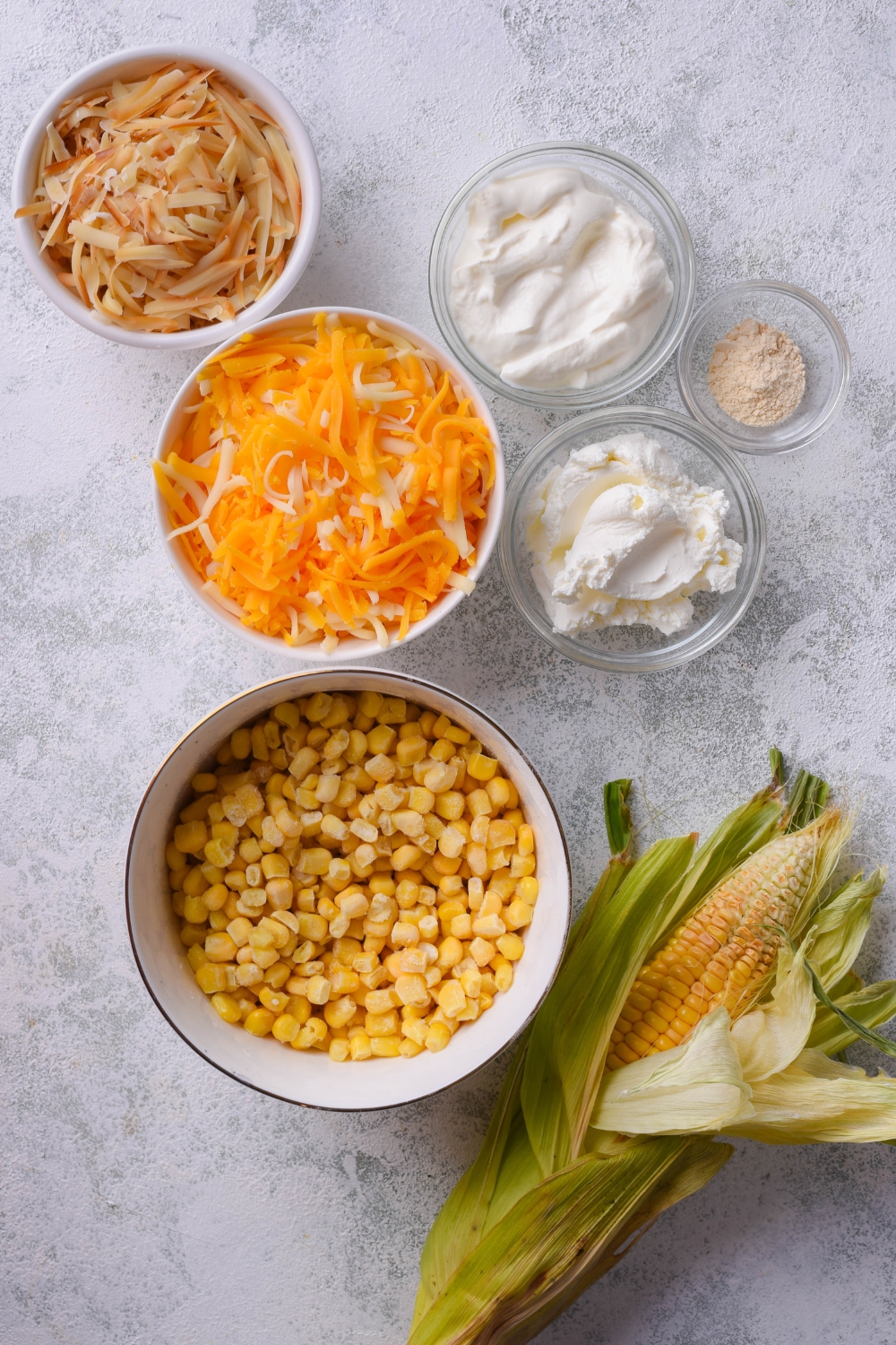 A countertop with separate bowls of shredded cheese, sour cream, cream cheese, salt and pepper, and frozen corn.