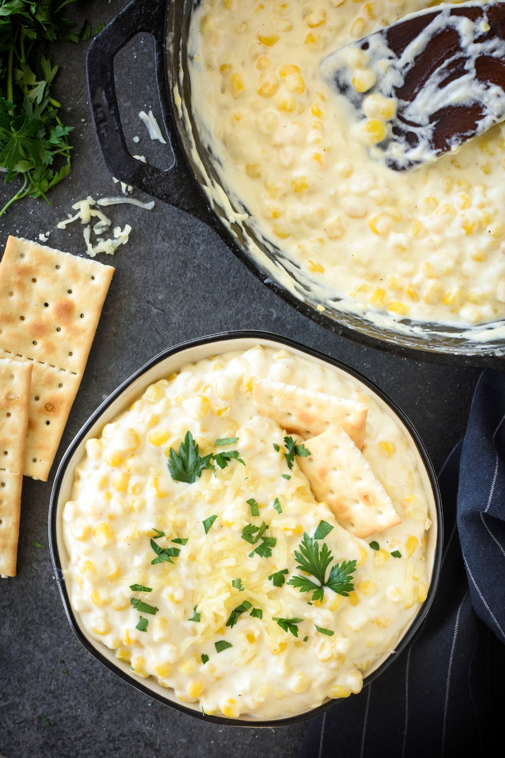 A skillet and bowl of cream cheese corn. The bowl is garnished with crackers and parsley and there's a wooden spatula in the skillet.