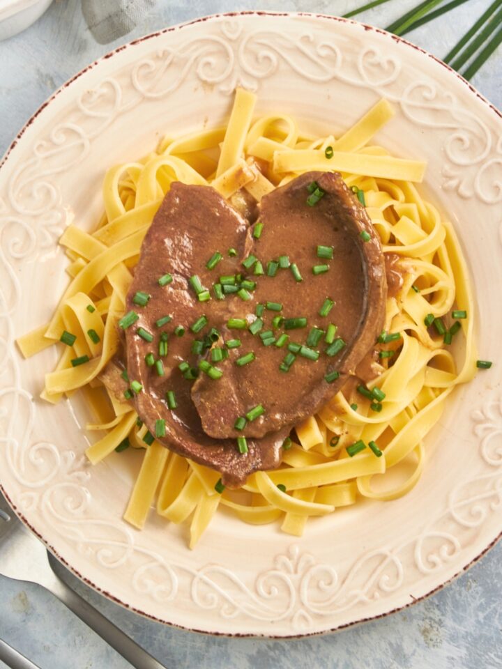 A piece of cubed steak with gravy on top of egg noodles on a white plate.