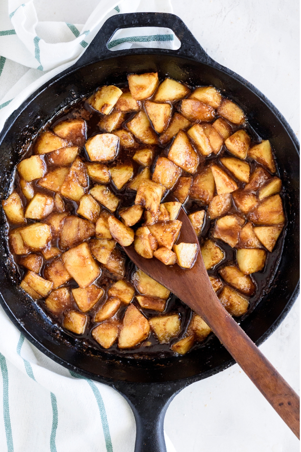 A cast iron skillet with fried apples and a wooden spoon stirring them.