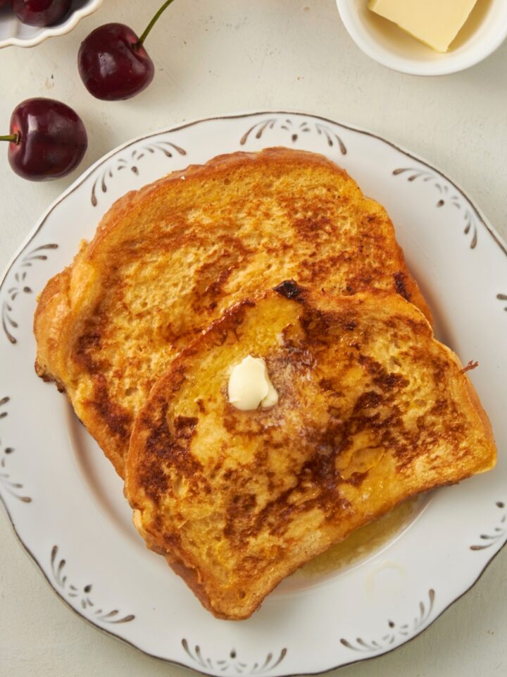 A square of butter on top of a slice of french toast that is on another slice of french toast on a white plate on a white counter.