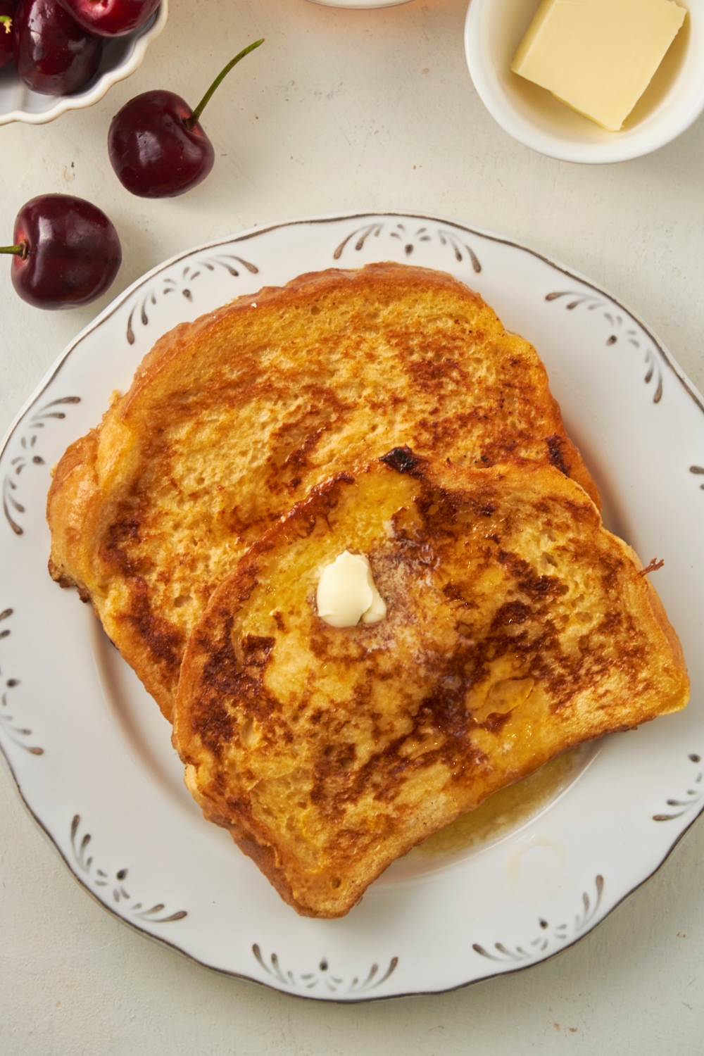 A square of butter on top of a slice of french toast that is on another slice of french toast on a white plate on a white counter.