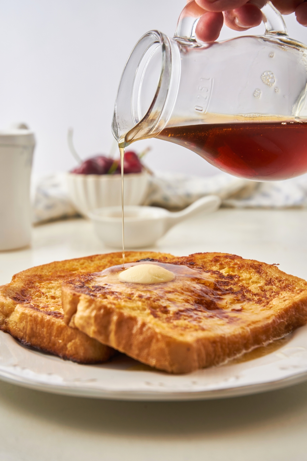 A hand pouring a pitcher of syrup on top of a slice of french toast on a white plate.