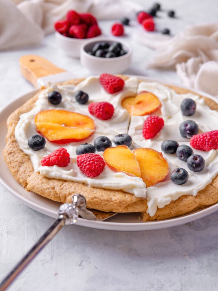 A sugar cookie fruit pizza on a white plate. It has a cream cheese frosting and sliced peaches, blueberries, and raspberries on top.