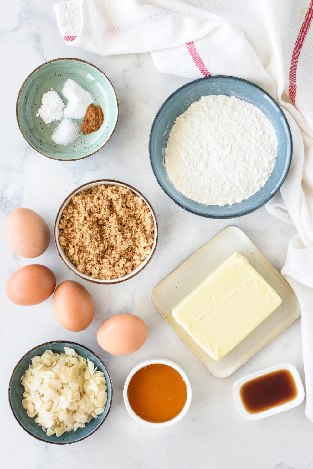 Bowls of flour, brown sugar, sliced almonds, and other dry ingredients, a plate of butter, eggs, and smaller bowls of honey and vanilla extract.