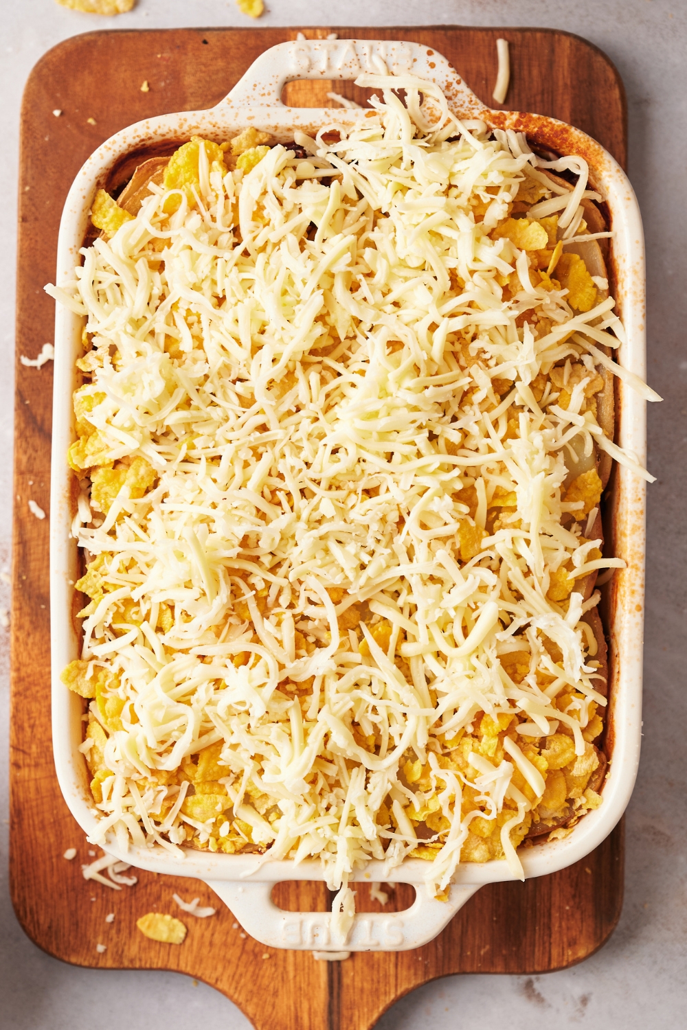 A casserole dish on a wooden serving board with baked shipwreck casserole with grated cheese and crushed cornflakes on top.