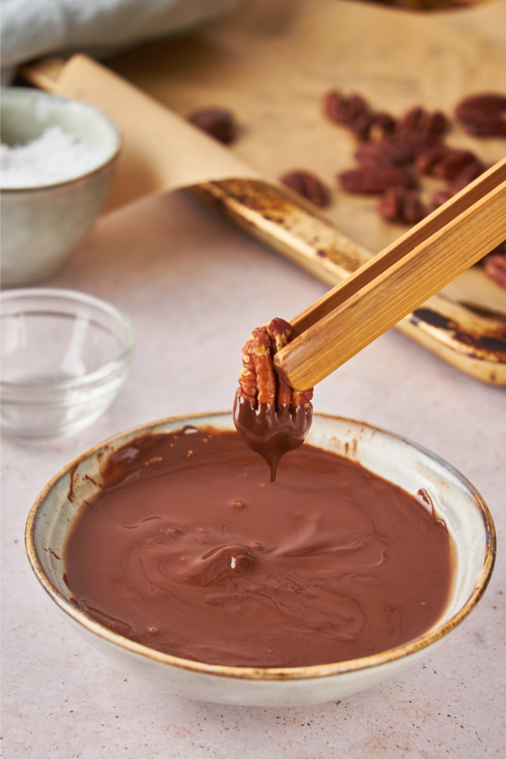 A small bowl with melted chocolate and a pair of tongs dipping individual pecans into the chocolate.