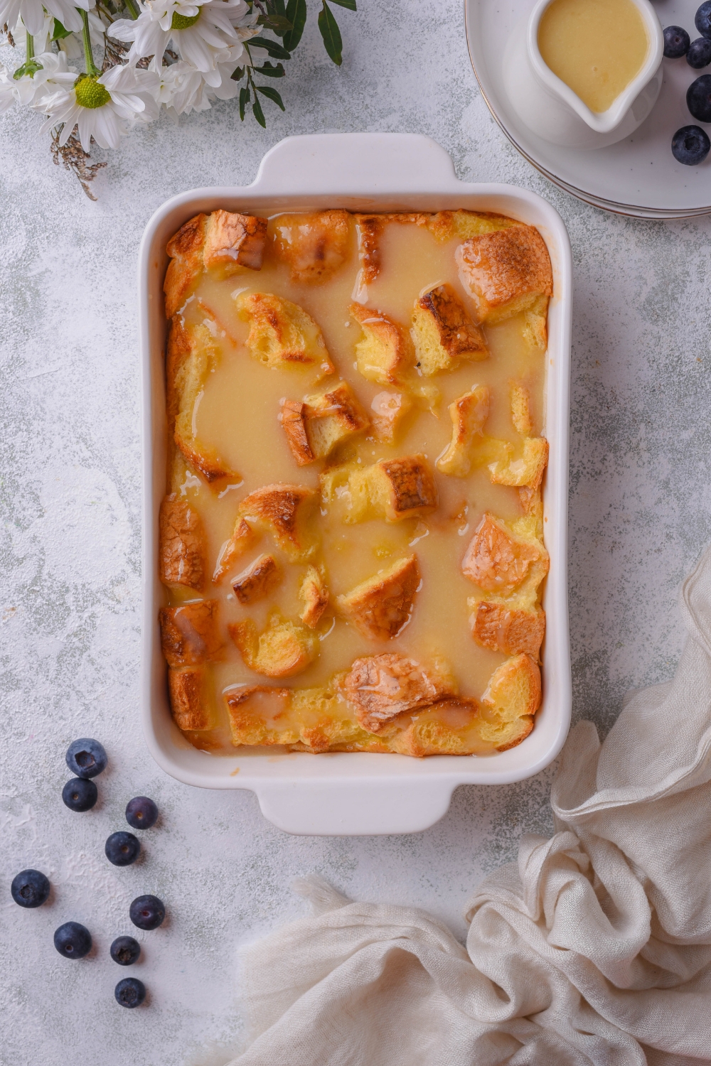 Bread pudding in a white baking tray, part of a pitcher of vanilla sauce is on the side.