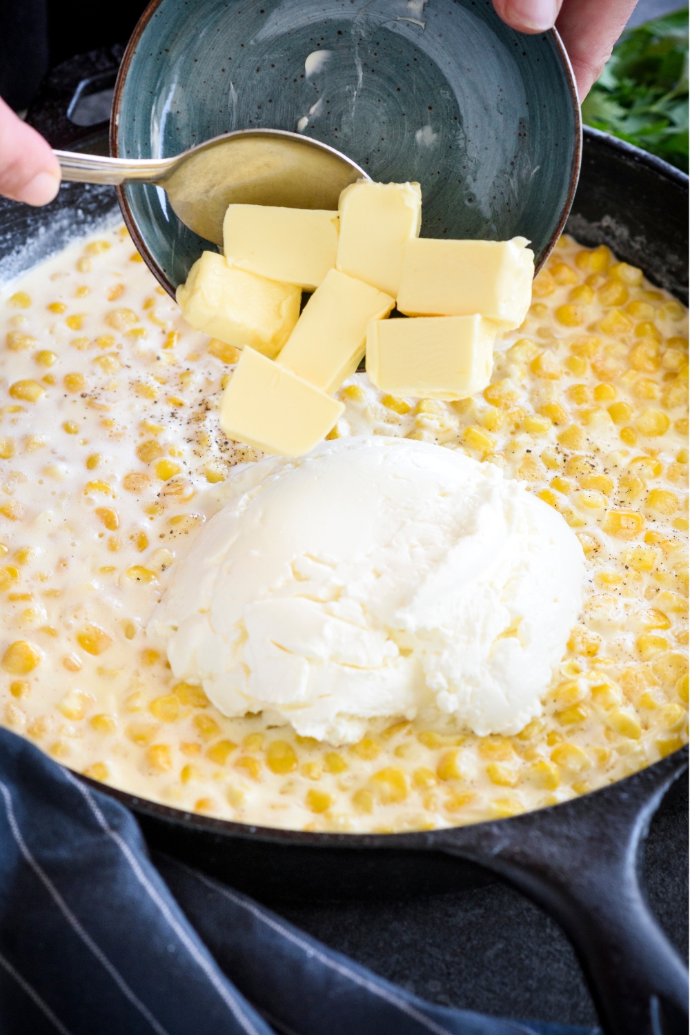 Knobs of butter being added to a skillet of corn, cream, and cream cheese.
