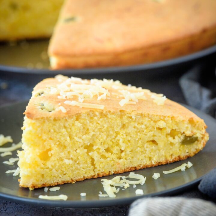 A slice of Jiffy jalapeno cornbread on a black plate. Behind it is the entire cornbread with the one slice missing from it.