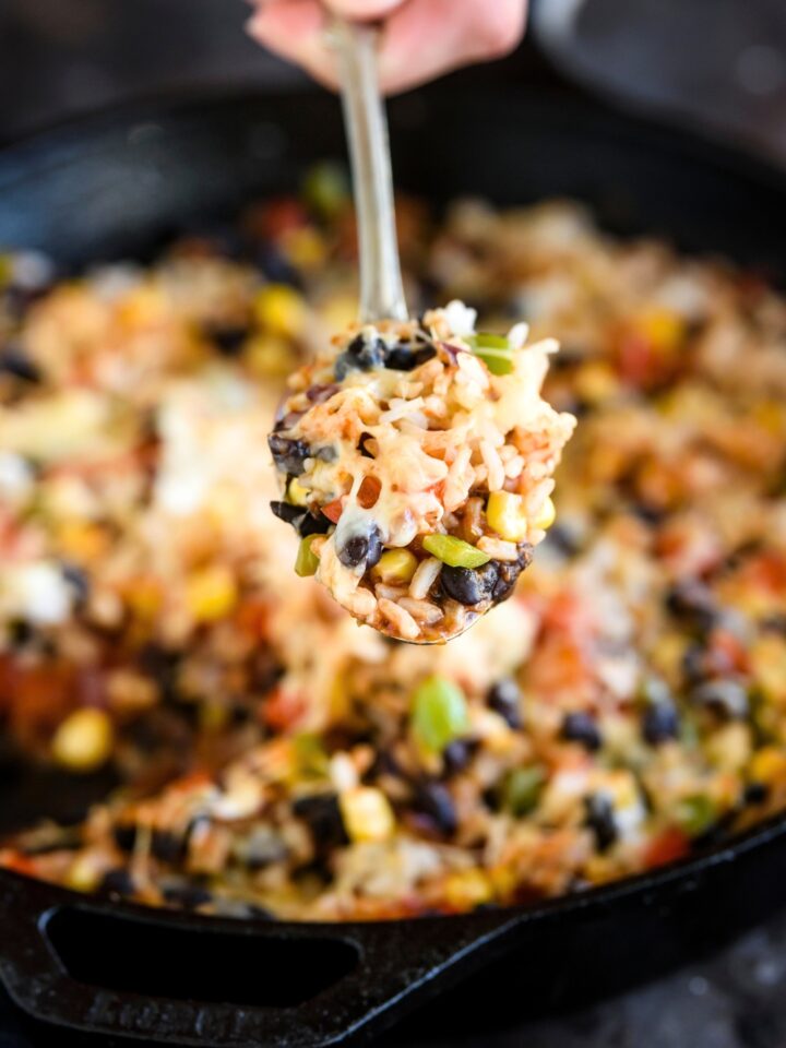 A hand holding a spoon that has Mexican rice casserole on it. Behind the spoon is a skillet filled with the casserole.