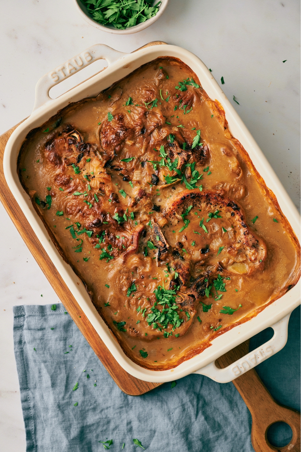 Four pork chops in a creamy soup in a white baking dish on top of a wooden cutting board on a white counter.