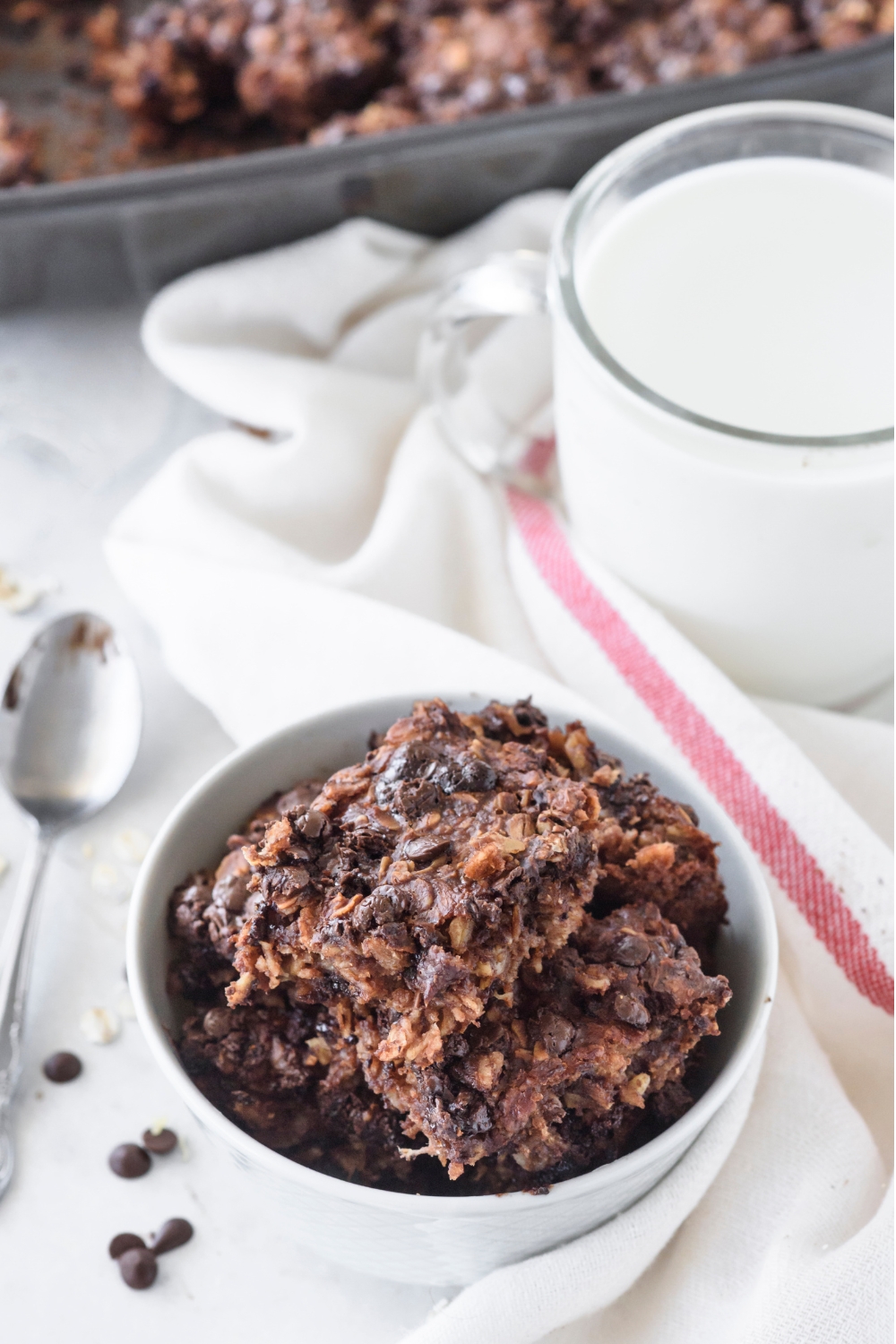 A bowl with a heaping pile of brownie baked oatmeal. A cup of milk and the remaining baked oats are in the background.