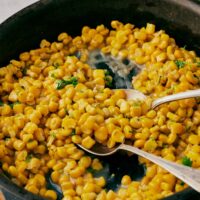 A cast iron skillet with fried corn topped with chopped cilantro. Two spoons are scooping the corn.
