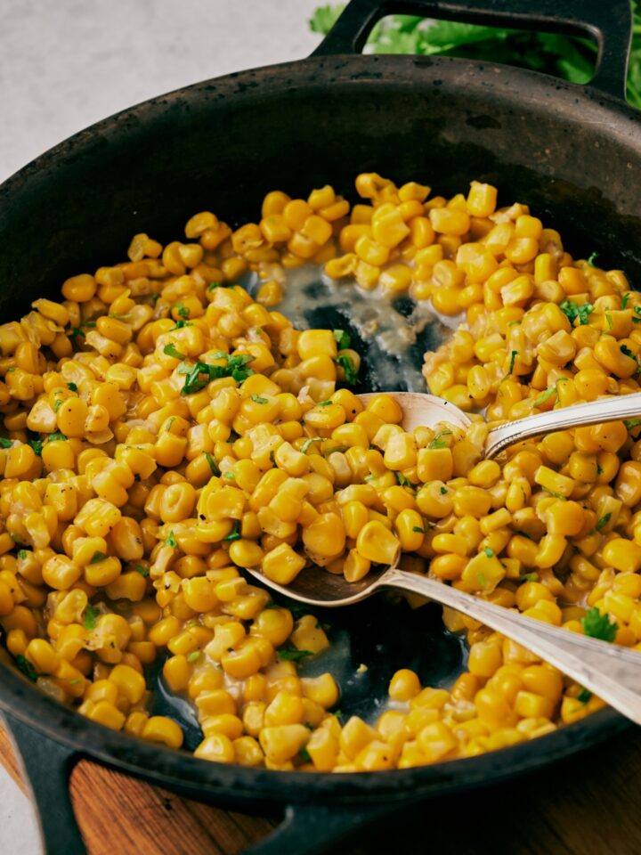 A cast iron skillet with fried corn topped with chopped cilantro. Two spoons are scooping the corn.