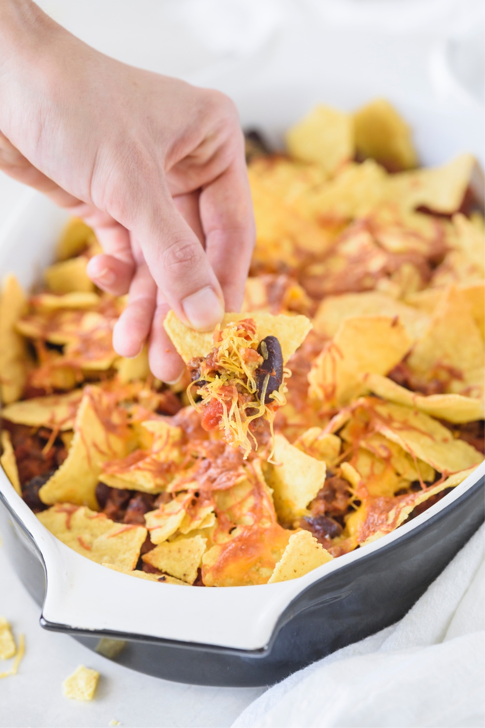 A baking pan with taco casserole a hand is holding a chip piled with taco casserole on it.