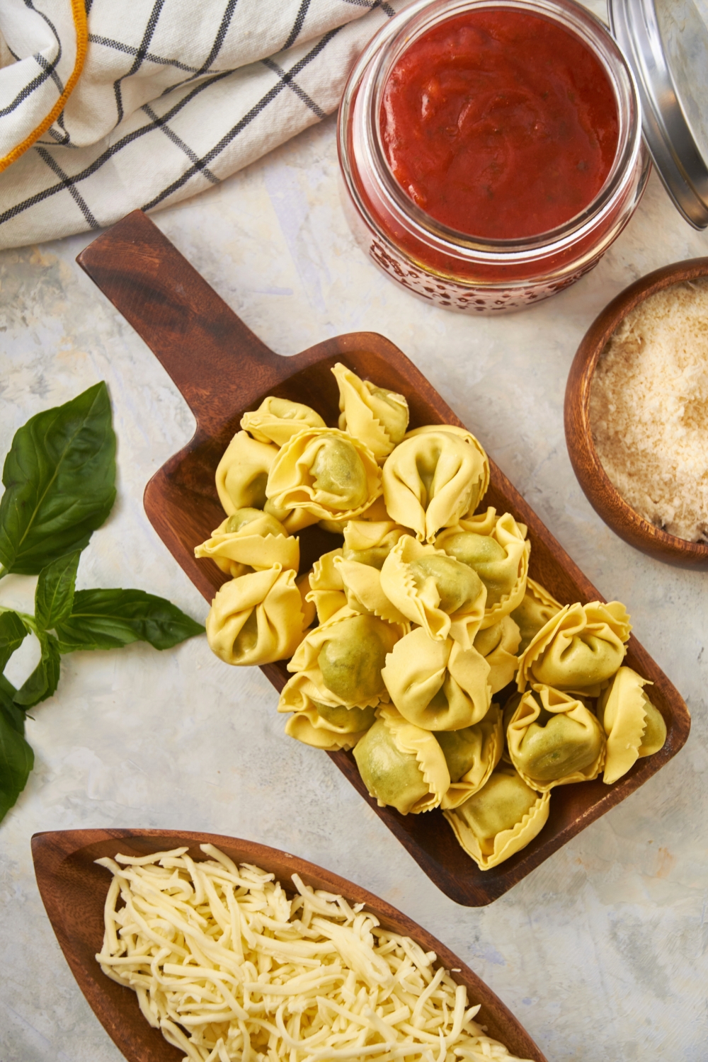 A bunch of tortellini on a wooden cutting board on a white counter. Surrounding it is a jar of pasta sauce and half a bowl of shredded mozzarella cheese.