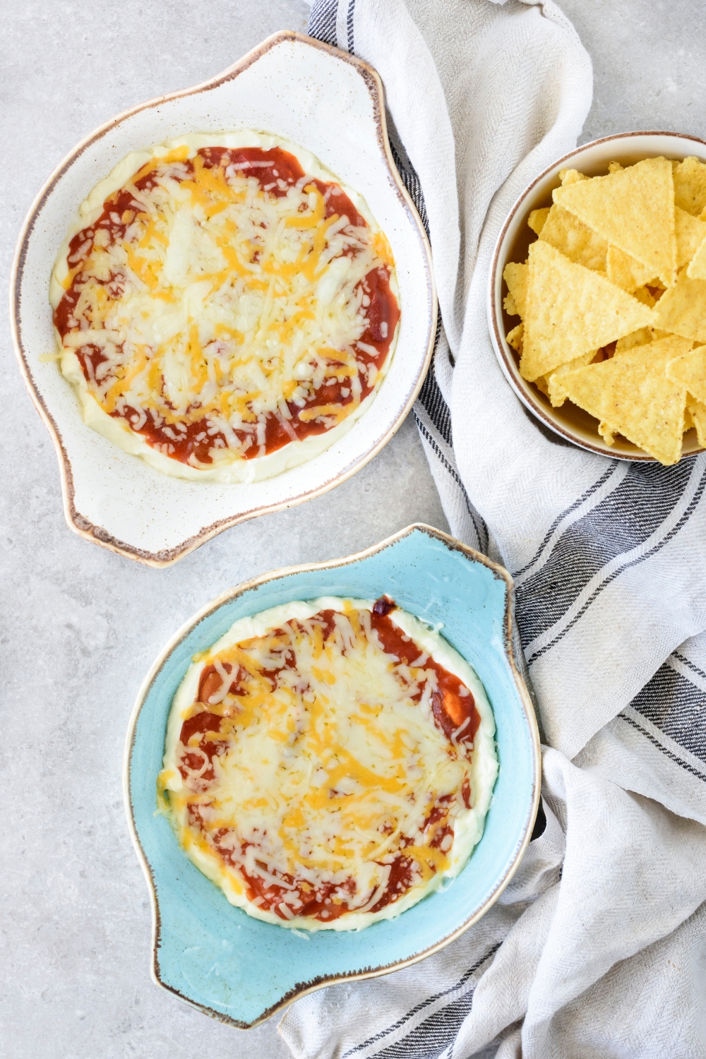 Two baking dishes filled with freshly baked chili cheese dip covered in melted cheese. There is a bowl of corn chips next to the bowls of dip.
