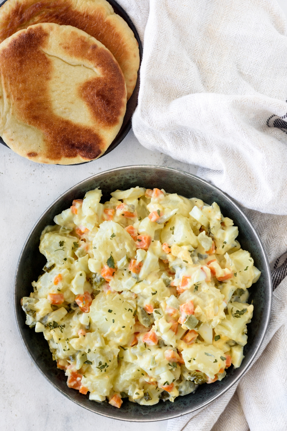A bowl of potato salad with chunks of carrot and pickles. Next to the bowl is a stack of two pita bread.