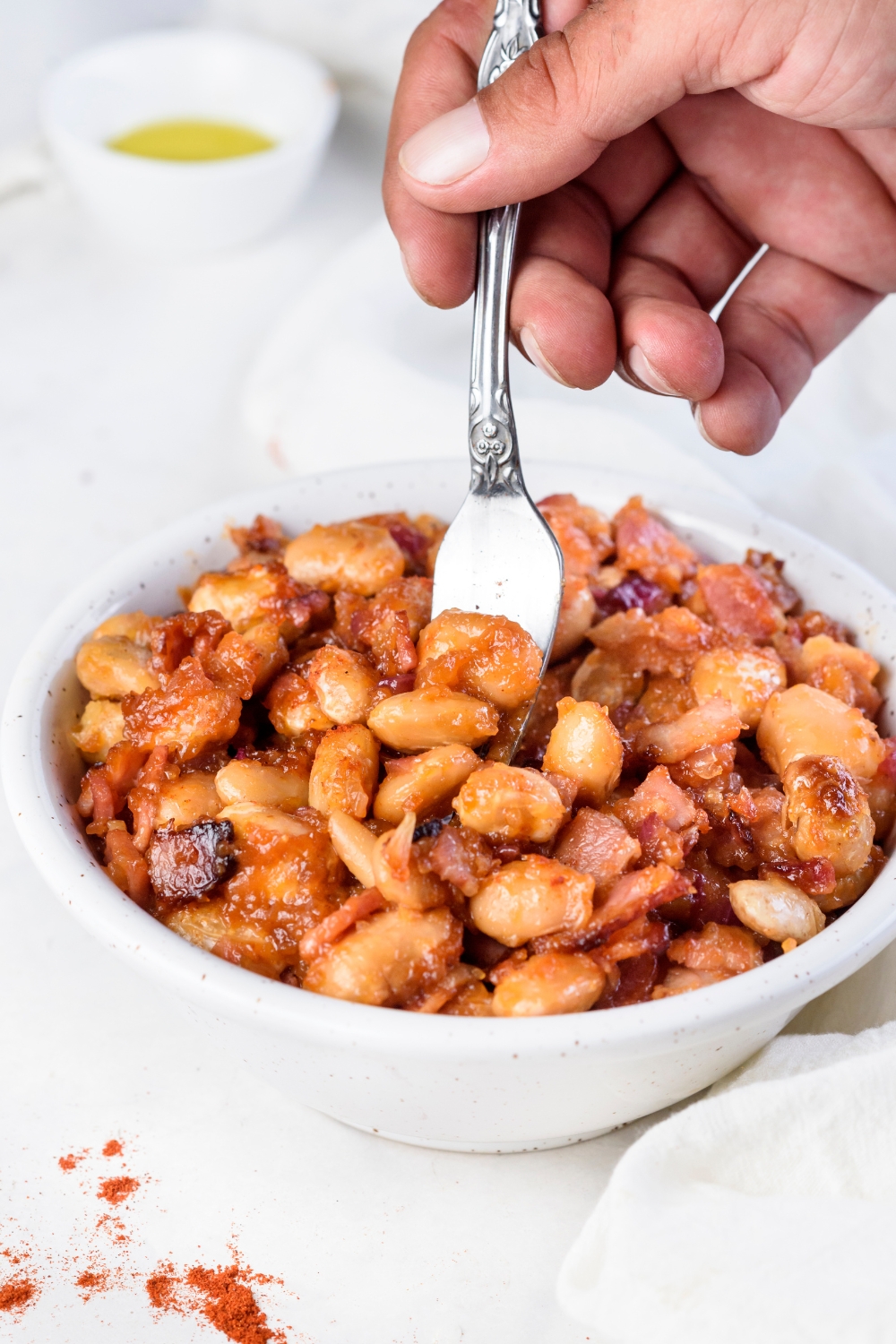 A hand using a fork to take a bite of baked beans out of a bowl filled with baked beans and bacon.
