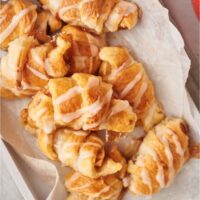 A pile of apple turnovers on a tray lined with parchment paper. They turnovers are drizzled with a sugar glaze.