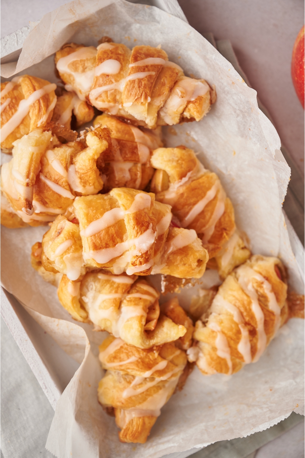 A pile of apple turnovers on a tray lined with parchment paper. They turnovers are drizzled with a sugar glaze.