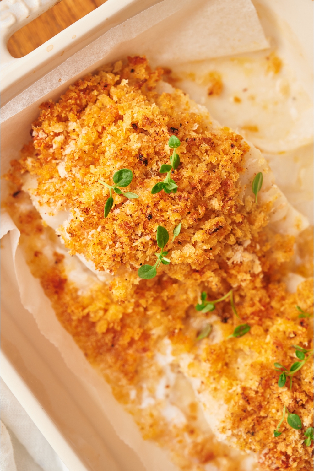 A baking dish with two cod fillets topped with a seasoned breadcrumb crust and garnished with fresh herbs.