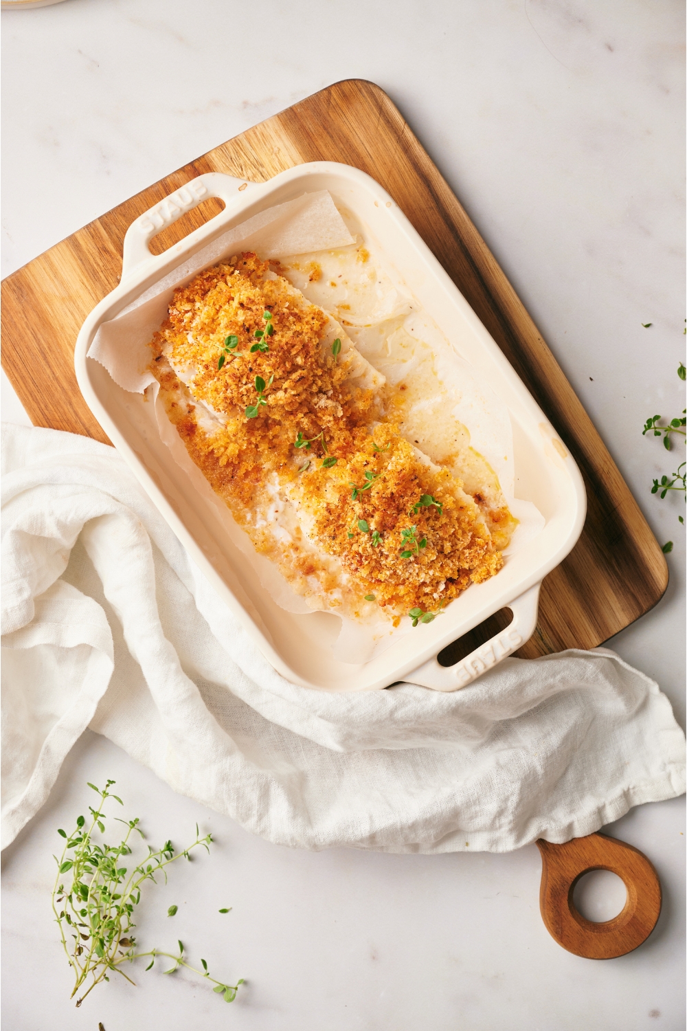 A baking dish with two cod fillets topped with a seasoned breadcrumb crust and garnished with fresh herbs.