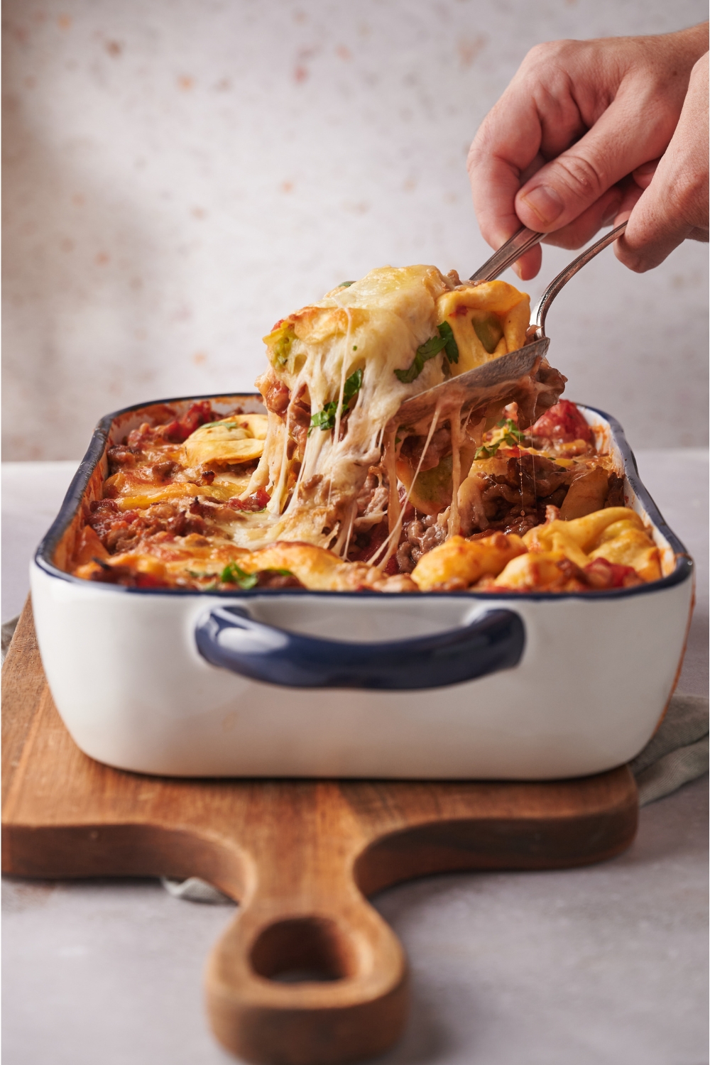 A person using two spoons to remove a serving of tortellini casserole from a baking dish. Cheese is stringing from the pasta.