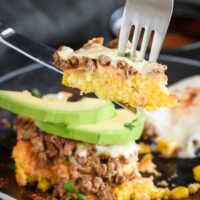 A fork and knife holding up a piece of beef tamale casserole with melted cheese and chunks of ground beef.