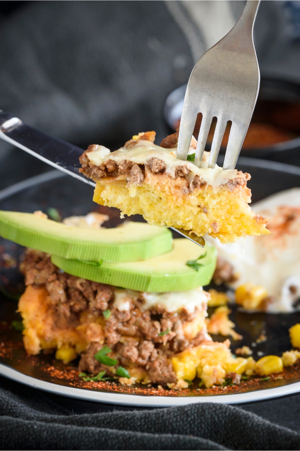 A fork and knife holding up a piece of beef tamale casserole with melted cheese and chunks of ground beef.