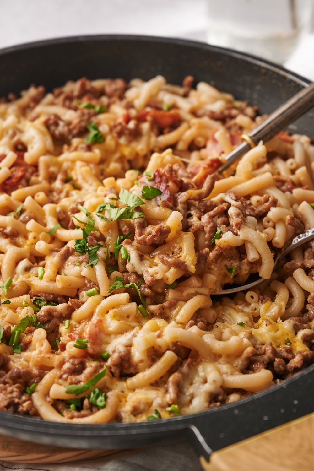 A skillet filled with bacon cheeseburger casserole with chunks of bacon and ground beef. The casserole is garnished with fresh herbs and there are two spoons in the skillet.
