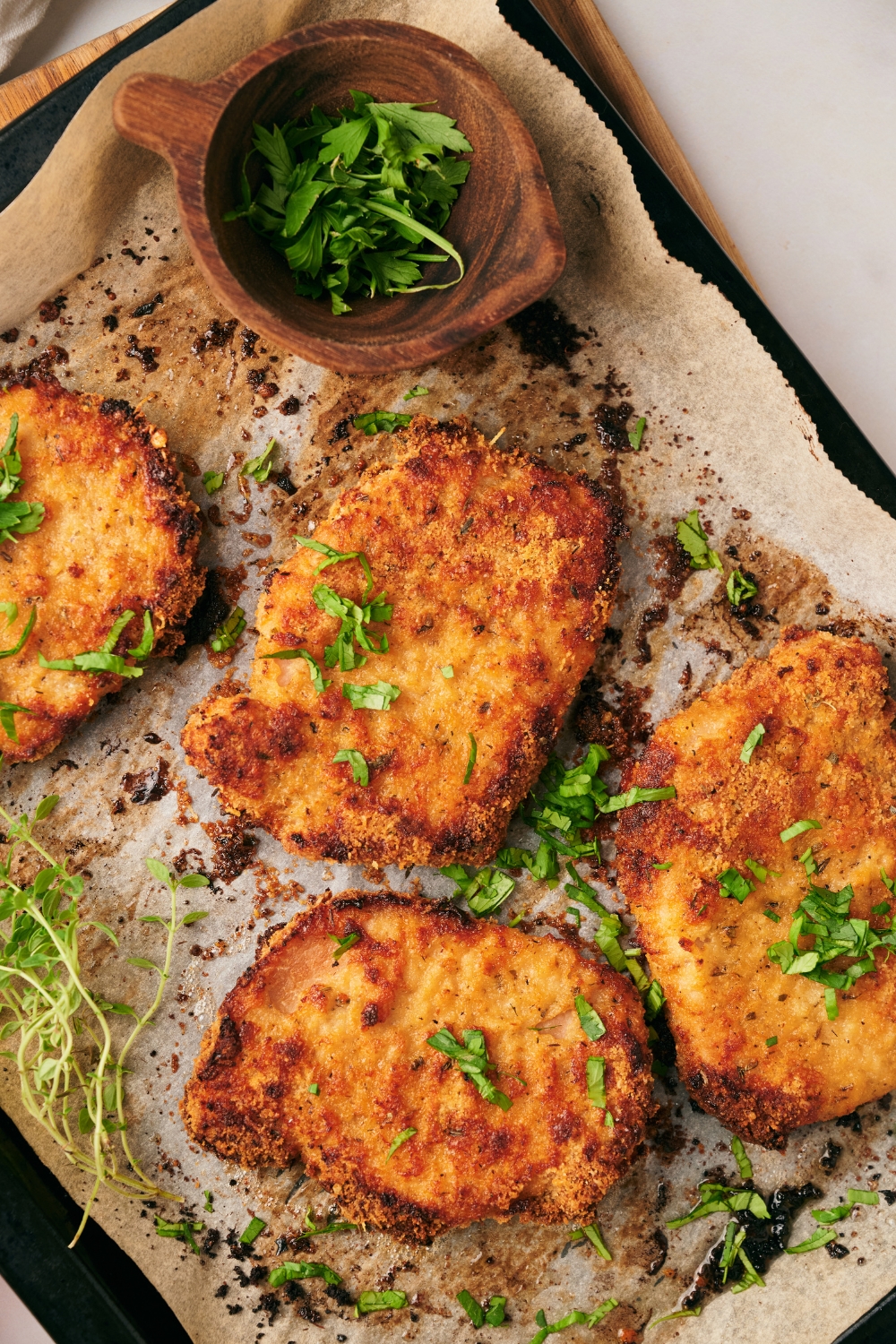 Four freshly baked and breaded pork chops on a baking sheet lined with parchment paper. Each is garnished with fresh green herbs.