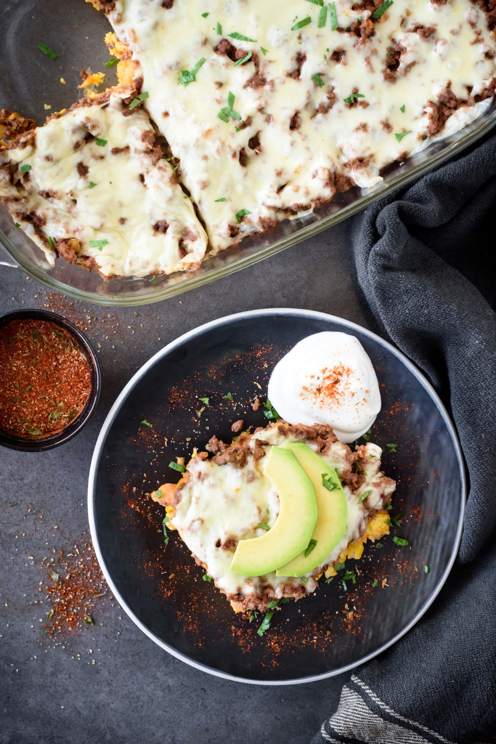 A serving of beef tamale casserole on a plate next to the rest of the casserole. The casserole is garnished with spices, sliced avocado, and sour cream.