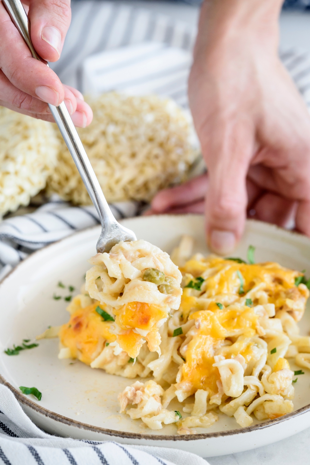 A hand using a fork to scoop a bite of cheesy tuna noodle casserole from a plate.