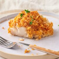 A cod fillet topped with a golden brown bread crumb crust and garnished with fresh herbs. There is a fork on the plate.